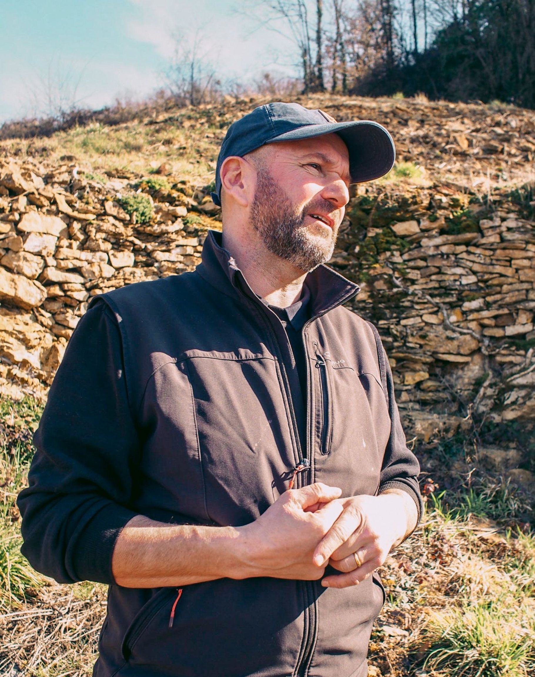 Winemaker of Tissot in a vest and cap, hands clasped. Stone wall and grassy hillside in background.