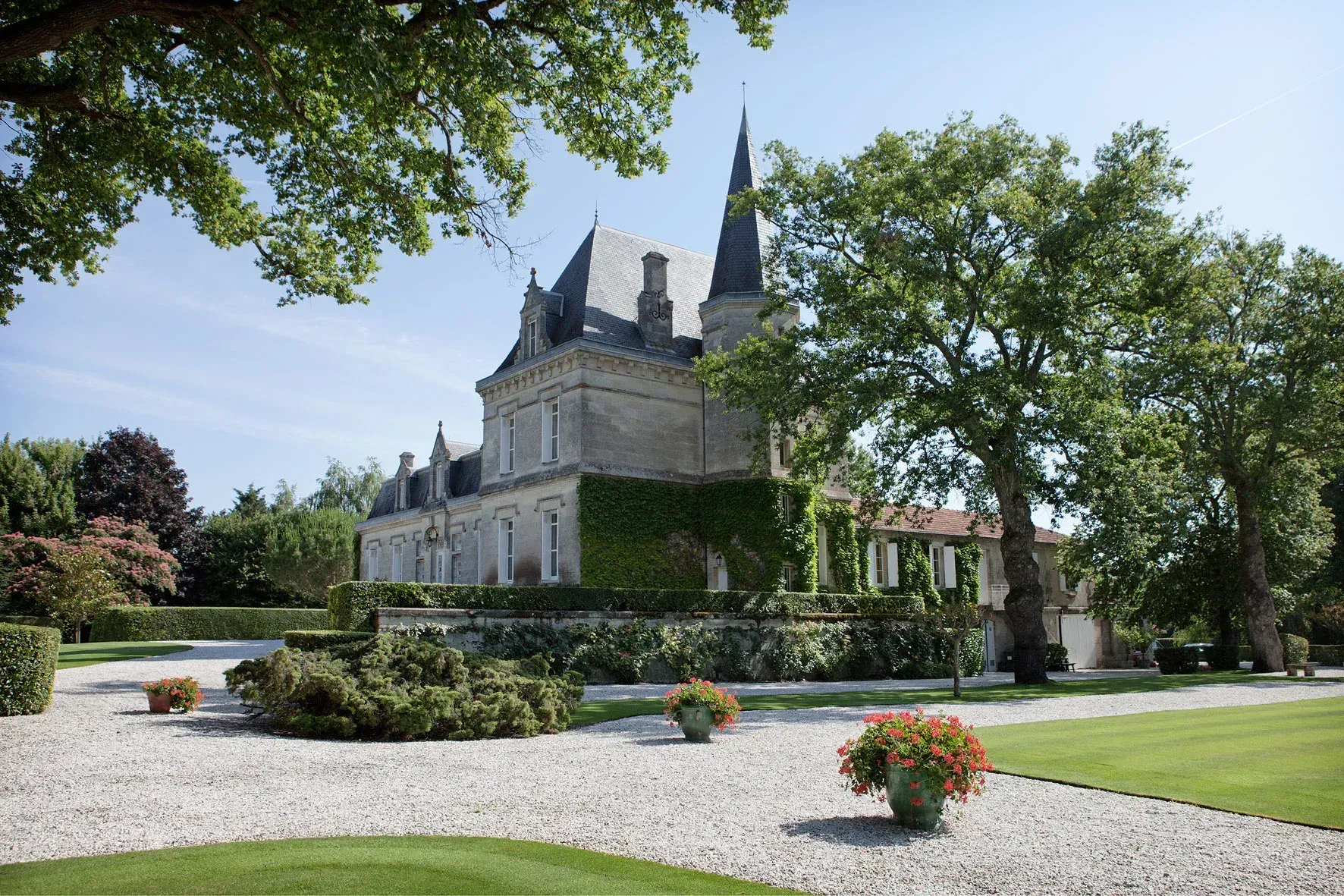 Stone chateau with a tall spire, partly covered in ivy, set in a manicured garden with gravel paths.