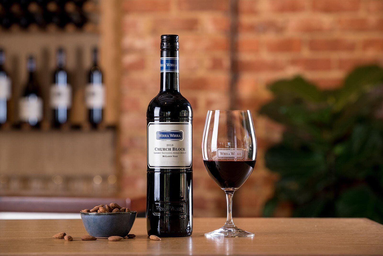 A bottle of Wirra Wirra Church Block beside a glass of red wine and a bowl of almonds.