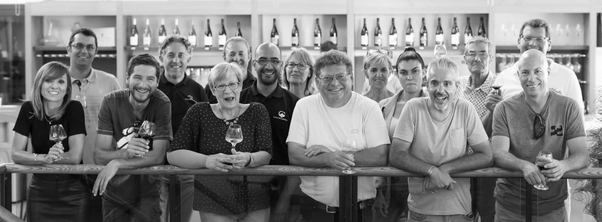 Cave de Cairanne team smiling,and holding wine glasses, with bottles on shelves in background.