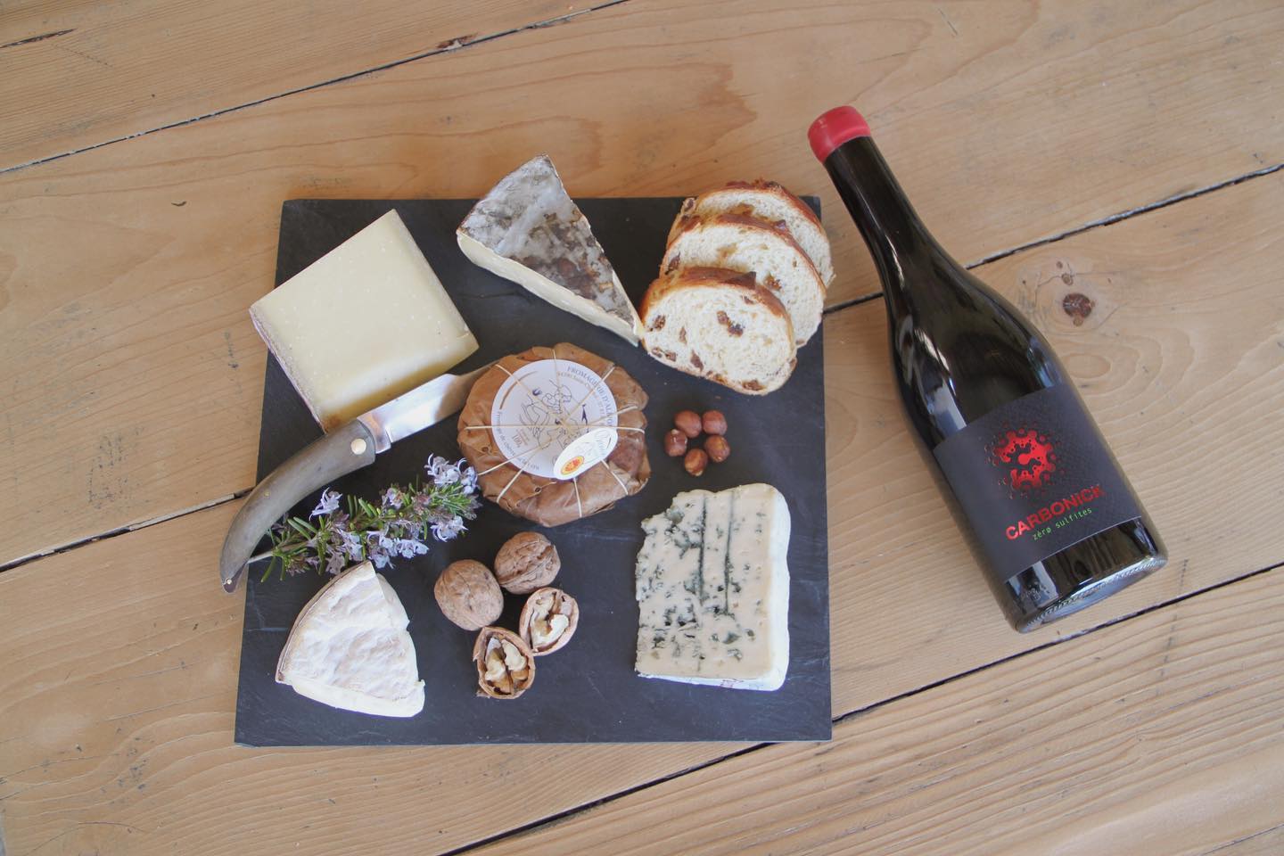 Cheese board with various cheeses, nuts, bread, and a bottle ofDomaine des Masques red wine on a wooden table.