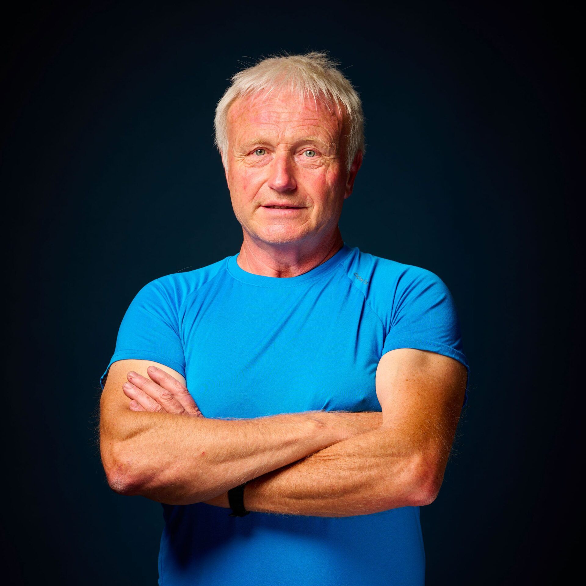 J. L. Denois on a blue shirt with arms crossed, on a dark blue background.