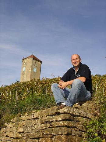 Tissot winemaker sitting on stone wall in vineyard, tower in background.