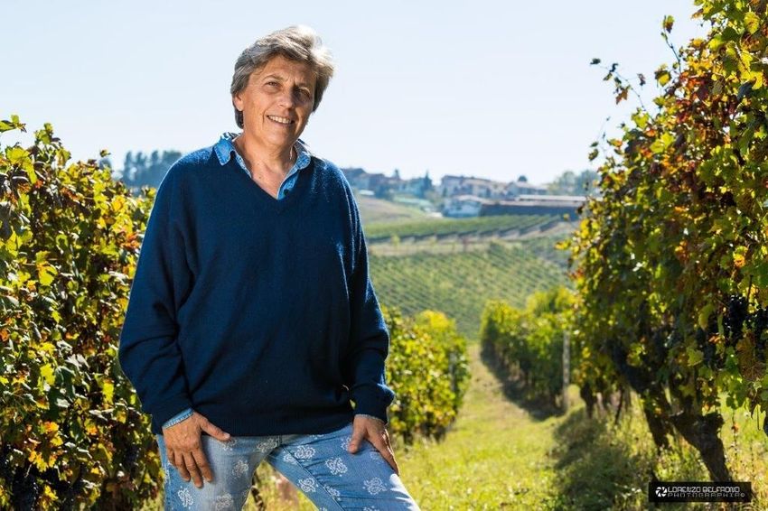 A winemaker smiling among autumn vines, the vineyard rows and a hilltop village behind her.